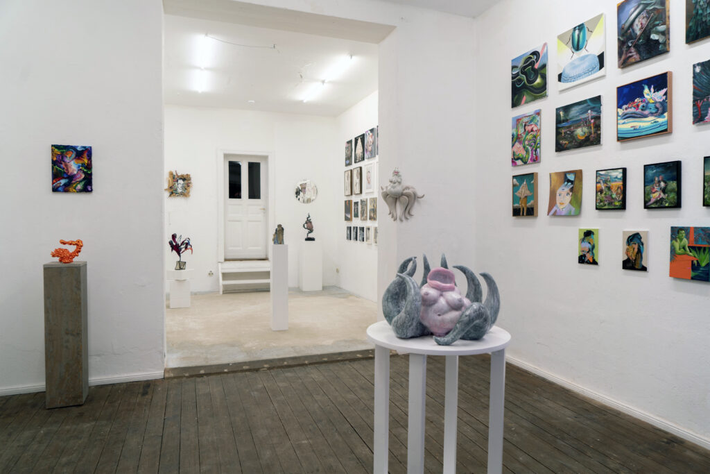 Exhibition view of Lusus Naturae at BMCA Berlin.in the foreground sits an octopus woman, Octomama on a round wooden base. her tentacles stretch groping upwards. in the background at the corner of a wall hangs the ceramic Big Sister, her tentacles are fanned out along the corner of the wall. both ceramics are lacquered and about 65x 60x 40cm large. 