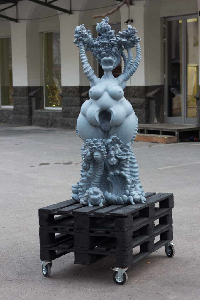 Eine göttin erhebt sich aus einem sockel aus wild wuchernden Pflanzen- Tier- und Frauendarstelungen. Ihre Arme werden zu ornamentalen Ästen, die sich mit den abstrahierten Haaren zu einer art Baumkrone vereinen.Die Figur ist blaugrau patiniert und 180cm hoch. sie steht auf einem schwarzen Palettenwagen