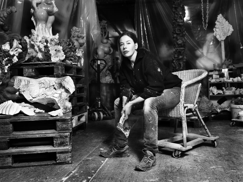 miriamlenk sits sideways on a chair in her berlin studio.with a plaster axe in her hand.in the background female figures, pallets and working material